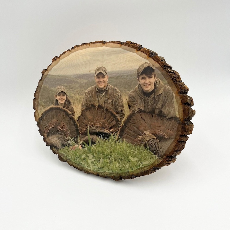 May include: A circular wood slice plaque featuring a printed photograph of three individuals in camouflage clothing posing with three wild turkeys in a grassy field. The natural bark edge remains visible around the perimeter of the wood piece.