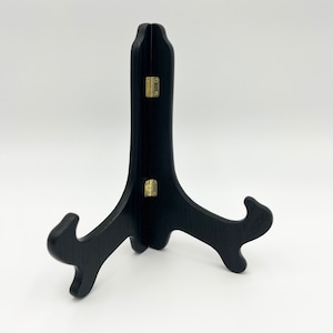 May include: A black wooden display stand with a visible wood grain texture. The stand features a hinged design connected by two small gold-colored metal hinges, allowing it to fold. It has a tall central support and two curved front arms.