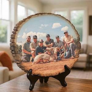 May include: A circular wood slice plaque featuring a printed photograph of five people posing with a bighorn sheep in a desert landscape. The plaque has natural bark edges and is displayed on a black plastic stand on a wooden surface.
