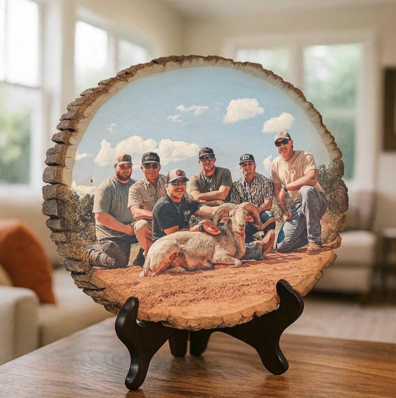 May include: A circular wood slice plaque featuring a printed photograph of five people posing with a bighorn sheep in a desert landscape. The plaque has natural bark edges and is displayed on a black plastic stand on a wooden surface.
