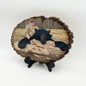 May include: A natural wood slice plaque featuring a printed photograph of a person in camouflage clothing sitting behind a large black bear resting its head on a fallen log. The plaque has a rough, bark-covered edge and rests on a black stand.