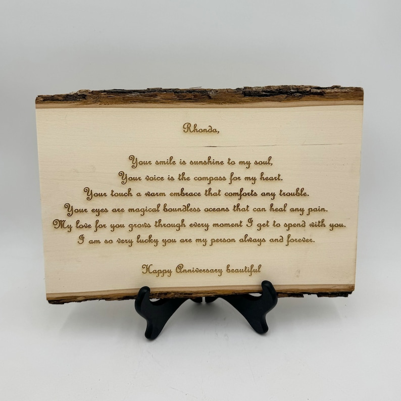 May include: A rectangular light-colored wooden plaque with natural bark edges, displayed on a black plastic stand. The surface features laser-engraved text: Rhonda, Your smile is sunshine to my soul, Your voice is the compass for my heart, Your touch a warm embrace that comforts any trouble. Your eyes are magical boundless oceans that can heal any pain. My love for you grows through every moment I get to spend with you. I am so very lucky you are my person always and forever. Happy Anniversary beautiful.