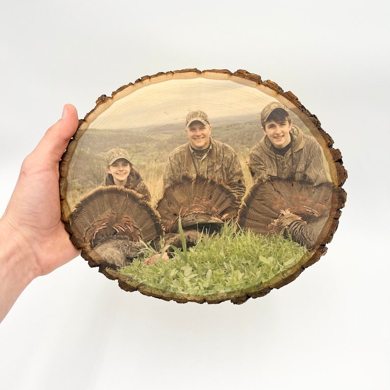 May include: A circular wood slice featuring a printed photograph of three individuals in camouflage clothing posing with three wild turkeys in a grassy field. The natural bark edge remains intact around the perimeter of the wooden plaque.