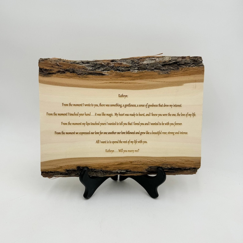 May include: A rectangular live-edge wood plaque featuring dark brown engraved text that reads: Kathryn: From the moment I wrote to you, there was something, a gentleness, a sense of goodness that drew my interest. From the moment I touched your hand... it was like magic. My heart was ready to burst, and I knew you were the one, the love of my life. From the moment my lips touched yours I wanted to tell you that I loved you and I wanted to be with you forever. From the moment we expressed our love for one another our love billowed and grew like a beautiful rose; strong and intense. All I want is to spend the rest of my life with you. Kathryn... Will you marry me? The plaque rests on a black plastic stand.
