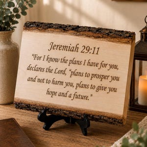 Peut inclure: Une plaque en bois rectangulaire avec des bords naturels sur le dessus et le dessous. La surface en bois clair est gravée avec le texte : Jeremiah 29:11, For I know the plans I have for you, declares the Lord, plans to prosper you.