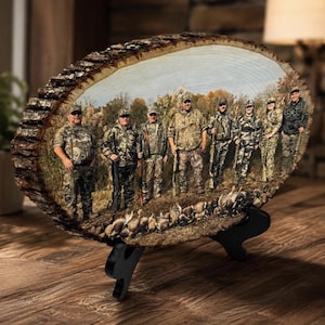 May include: A circular wood slice plaque featuring a printed photograph of eight individuals in camouflage hunting gear standing in a field with harvested game birds at their feet. The plaque has a natural bark edge and rests on a black stand.