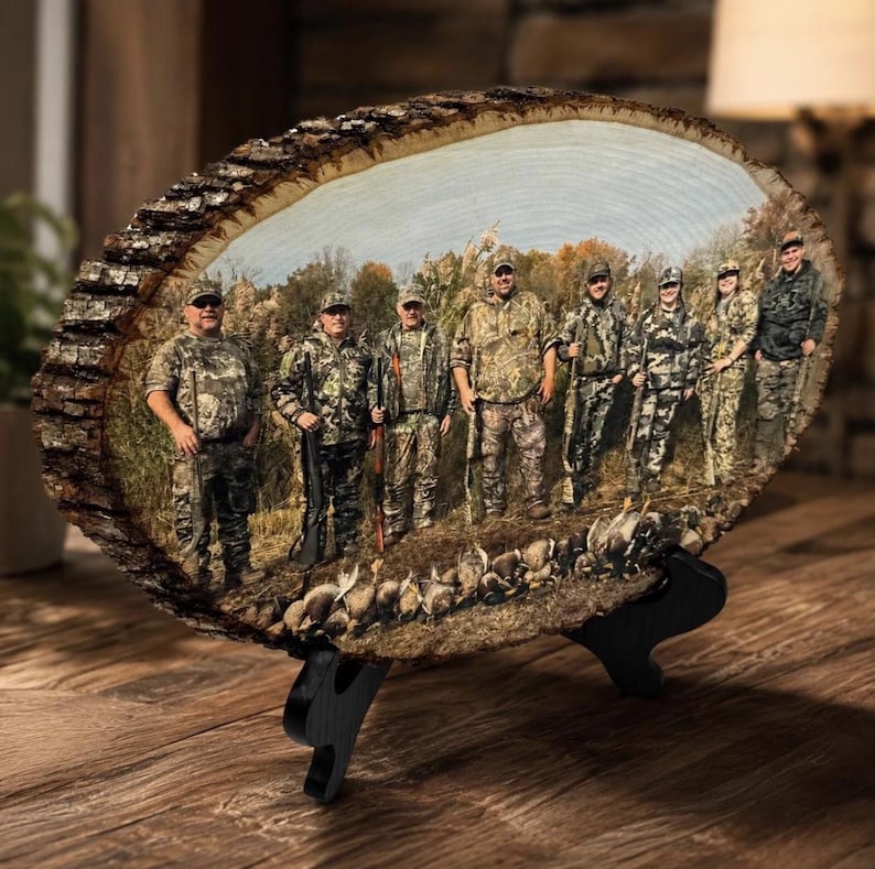 May include: A circular wood slice plaque featuring a printed photograph of eight individuals in camouflage hunting gear standing in a field with harvested game birds at their feet. The plaque has a natural bark edge and rests on a black stand.
