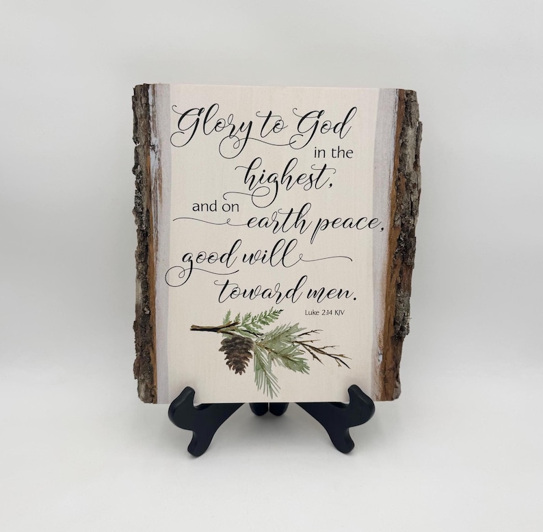 May include: A wooden sign with a white background and brown bark edges. The sign features a black calligraphy quote: "Glory to God in the highest, and on earth peace, good will toward men." The quote is from Luke 2:14 KJV. A watercolor painting of a pine branch with a pine cone is in the bottom right corner.