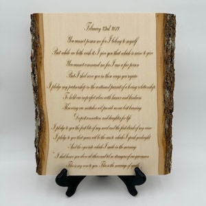 May include: A rectangular light-colored wooden plaque with natural bark edges on the left and right sides. The surface features dark engraved cursive text starting with February 23rd 2019, followed by a long wedding vow. It rests on a black stand.