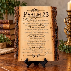 May include: A rectangular light-colored wooden plaque with natural dark bark edges, featuring the engraved text of Psalm 23. The plaque is supported by a black plastic stand and displays the full biblical passage in a mix of serif and script fonts.