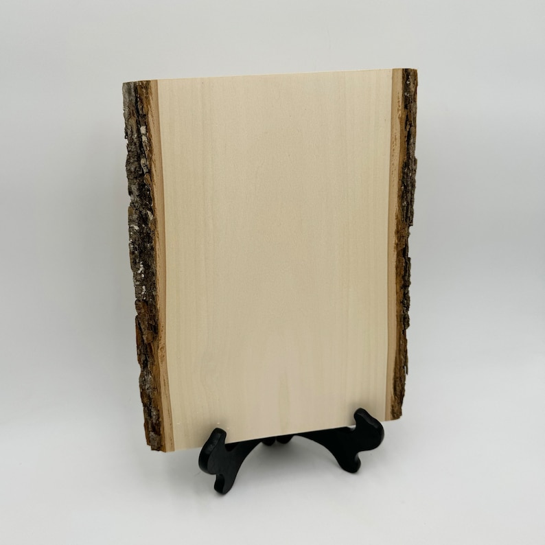 Puede incluir: Una rebanada de madera natural con un borde de corteza &aacute;spero, montada en un soporte de metal negro. La madera es de color claro y tiene una superficie lisa.