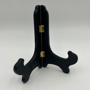 May include: A black wooden display stand featuring a two-piece folding design connected by two small gold-toned metal hinges. The stand has a curved, upright support structure with two outward-curving arms at the base to hold items securely in place.