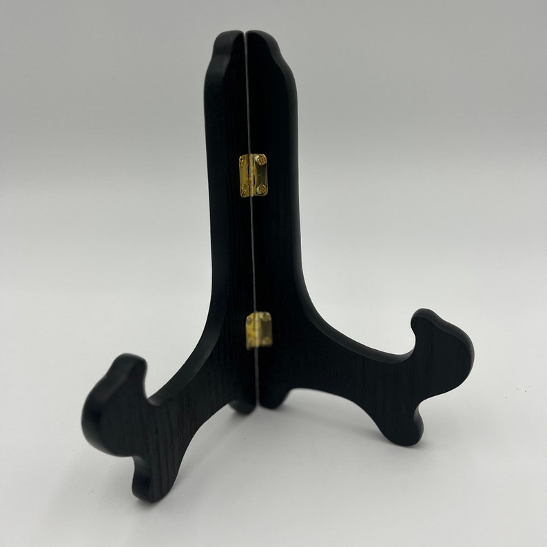 May include: A black wooden display stand featuring a two-piece folding design connected by two small gold-toned metal hinges. The stand has a curved, upright support structure with two outward-curving arms at the base to hold items securely in place.