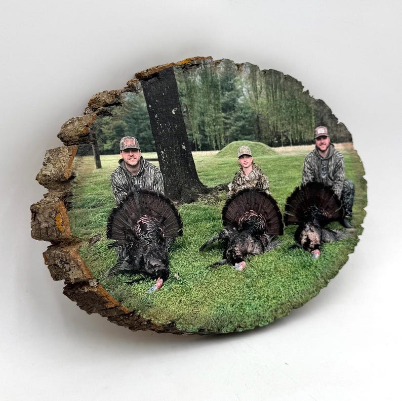 May include: A circular wood slice plaque featuring a printed photograph of three individuals in camouflage clothing posing in a grassy field with three wild turkeys. The natural bark edges of the wood slice frame the outdoor hunting scene.