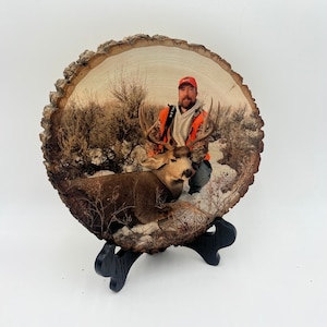 May include: A circular wood slice featuring a printed photograph of a person in a bright orange hunting vest and cap kneeling beside a large buck with antlers in a snowy, brush-filled landscape. The wood slice rests on a black display stand.