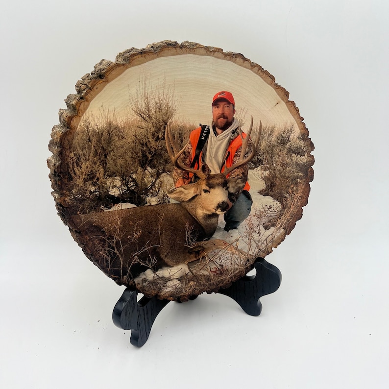 May include: A circular wood slice featuring a printed photograph of a person in a bright orange hunting vest and cap kneeling beside a large buck with antlers in a snowy, brush-filled landscape. The wood slice rests on a black display stand.