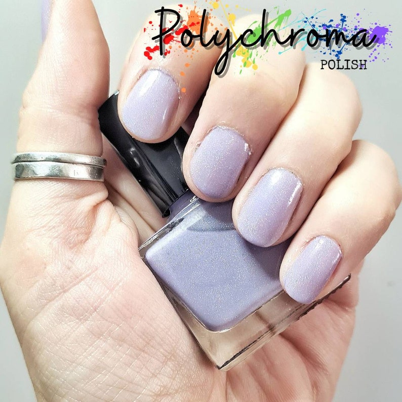 May include: A hand model wearing a silver ring, showcasing a bottle of light purple nail polish with a shimmer finish. The bottle is labeled "Polychroma POLISH".