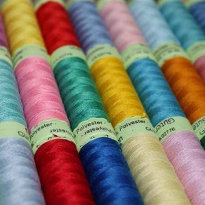 May include: A close-up of a selection of spools of thread in various colours, including red, blue, green, yellow, and pink. The spools are labelled with the word "Polyester" and the brand name "Gutermann".