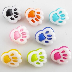May include: Assortment of ten colourful plastic paw print buttons. The buttons are white with a paw print design in various colours, including black, orange, blue, pink, yellow, and green.