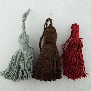 May include: Three decorative tassels in different colours: light blue, brown, and red. The tassels are made of soft, fluffy yarn and have a knotted top.