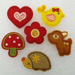 May include: Six felt appliques in various shapes and colours, including a red heart, a yellow bird, a red flower, a brown deer, a red mushroom, and a brown hedgehog.