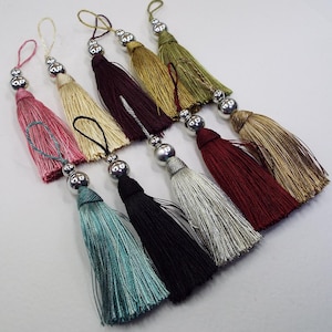 May include: Assortment of ten colourful tassel ornaments with silver metal beads. The tassels are made of soft, silky fabric and come in a variety of colours, including pink, beige, burgundy, gold, and silver.