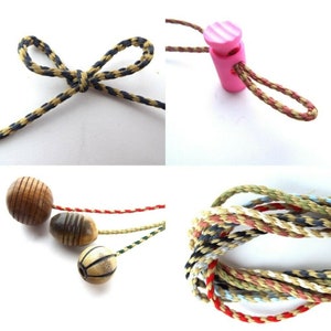 May include: Assortment of cords and beads for crafting projects. The cords are in various colours and patterns, including black and gold, tan and black, and red, white, blue, and tan. The beads are wooden and include a round brown bead, a striped brown bead, and a round bead with a basketball pattern.