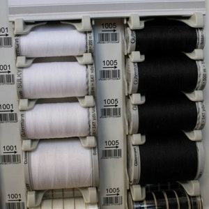 May include: A close-up of a display of white and black bobbins of thread. The bobbins are labelled with the number 1005 and the brand name Gütermann. The bobbins are arranged in rows on a white background.