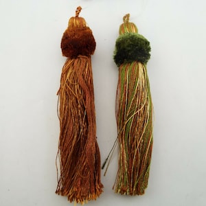 May include: Two decorative tassels, one brown and one green with gold accents. The tassels are made of a soft, fluffy material and have a long, flowing fringe.