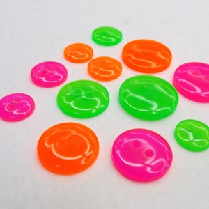 May include: A collection of brightly coloured plastic buttons in neon pink, green, and orange. The buttons have a smiley face design and two holes for sewing.