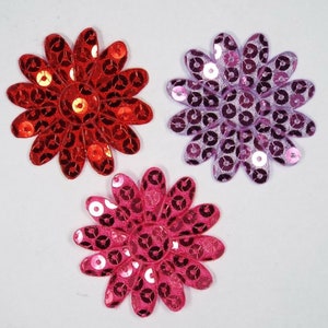 May include: Three decorative flower-shaped fabric patches with sequins. The patches are red, pink, and purple.