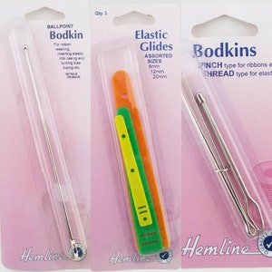 May include: Three packs of sewing tools. The first pack contains a silver ballpoint bodkin. The second pack contains three plastic elastic glides in orange, green, and yellow. The third pack contains two silver bodkins, one with a loop at the end. The packs are all pink and white with the Hemline logo.