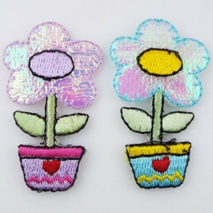 May include: Two embroidered iron-on patches of flowers in pots. One flower is light blue with a yellow centre and the other is purple with a purple centre. Both flowers have green leaves and are in pots with red hearts.