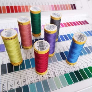 May include: A close-up of ten spools of thread in various colours, including red, green, blue, yellow, and pink. The spools are arranged on a white surface with a colour chart in the background. The spools are labelled with the brand name "Gütermann" and the colour number.