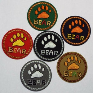 May include: Six embroidered iron-on patches featuring a bear paw print and the word "BEAR" in a circle. The patches are in various colours: green, navy blue, red, black, grey, and brown.
