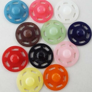 May include: Assortment of ten plastic snap buttons in various colours, including blue, pink, brown, green, navy, light pink, red, yellow, and orange. Each button has a circular shape with a raised centre and a decorative pattern of holes around the edge.