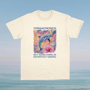 Op de afbeelding: Een licht beige t-shirt met een grafische print van een dolfijn die uit het water springt met een roze bloem. De tekst "Undiagnosed but something is definitely wrong" is onder de print gedrukt.
