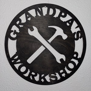 May include: A black circular wooden sign with the words "Grandpa's Workshop" written around the edge. A wrench and hammer are crossed in the center of the sign.