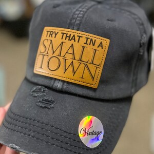 Puede incluir: Gorra de béisbol negra desgastada con un parche de cuero marrón que dice "Try that in a small town". La gorra tiene una pegatina vintage en el lateral.