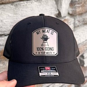 May include: Black trucker hat with a mesh back and a curved brim. The hat features a patch with the text "MY MEAT IS 100% GOING IN YOUR MOUTH" and a graphic of a grill. The hat is adjustable.