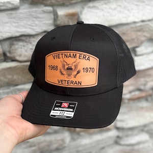 May include: Black trucker hat with a brown leather patch that says "Vietnam Era 1968-1970 Veteran". The hat has a black mesh back and a Richardson 112 adjustable strap.