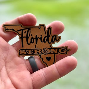 May include: A brown outline of the state of Florida with the text "Florida Strong" and a heart in the center. The outline is made of a textured material and has a black swirl design.