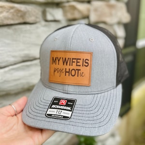 May include: Grey and black trucker hat featuring the text "MY WIFE IS psyc HOTic" on a brown leather patch. The hat has a curved peak and an adjustable strap. The brand tag reads "RICHARDSON ORIGINAL 112 TRUCKER".