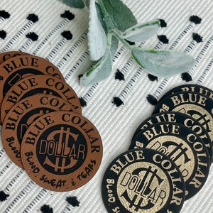 May include: Close-up of six round leather patches with the text "Blue Collar Dollar Blood Sweat & Tears" in black and gold lettering. The patches are arranged in two stacks of three, with the top patches in each stack facing the camera.