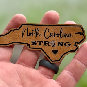 May include: A brown leather outline of the state of North Carolina with the text "North Carolina - STRONG" and a heart with a hurricane symbol inside.