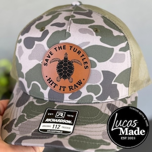 May include: Camouflage trucker hat with a brown leather patch featuring a turtle graphic and the text "Save the Turtles. Hit it Raw." The hat has a green mesh back and a curved brim. The Richardson brand label is visible.