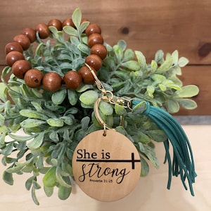 Può includere: Portachiavi in legno con un braccialetto di perline di legno marrone attaccato. Il portachiavi presenta un'etichetta rotonda in legno con il testo "She is Strong" e "Proverbi 31:25" in lettere nere. Una piccola croce dorata è accanto al testo. Il portachiavi ha anche un fiocco turchese.