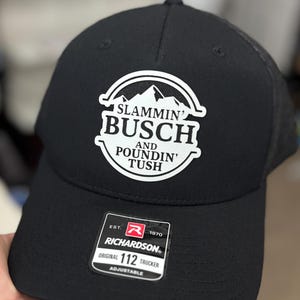 May include: Black trucker hat with a white patch featuring the text "SLAMMIN' BUSCH AND POUNDIN' TUSH" above a mountain range graphic. The hat has a mesh back and an adjustable strap. The Richardson brand label is visible.