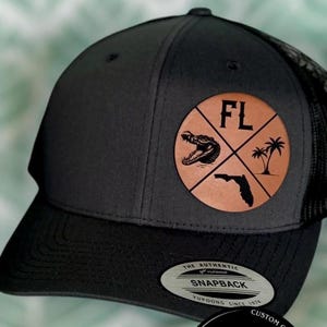 May include: A black and gray trucker hat with a leather patch featuring an alligator, palm trees, and the Florida outline. The patch has the letters "FL" and is on a gray crown and black brim.