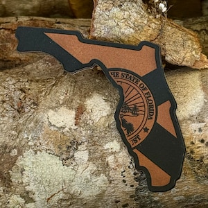 May include: A brown and black leather sticker of the state of Florida with the state seal in the center. The seal features a sun, a tree, and the words "The State of Florida".
